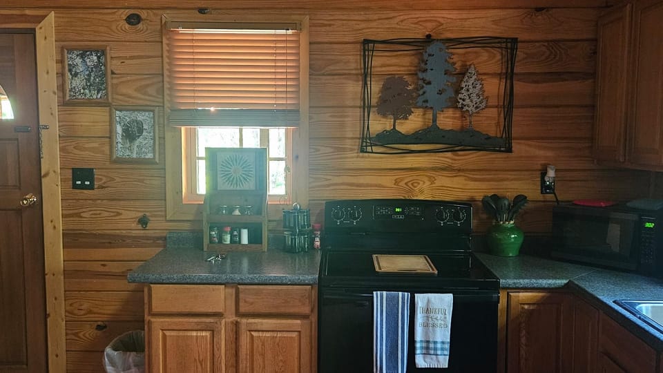 Compact kitchen with black electric stove, wood cabinets, granite countertops, and a window with blinds.