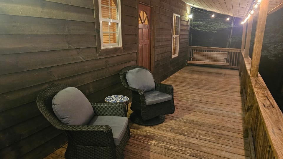 Wood-paneled porch with two cushioned black wicker chairs, ceramic side table, string lights, and a wooden swing bench.