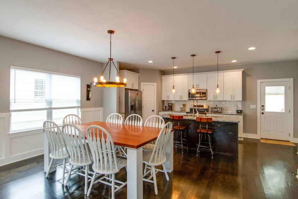 Dining area with a wood table, white chairs, pendant lights, and kitchen in background