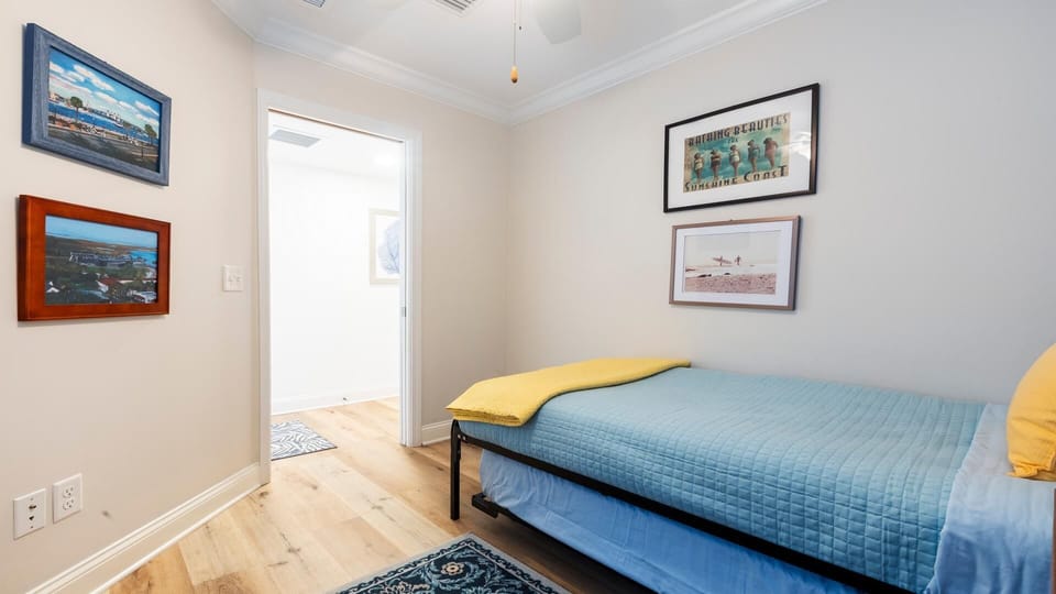 Single bed with a blue quilt and yellow throw in a bedroom featuring light wood flooring and framed beach artwork.