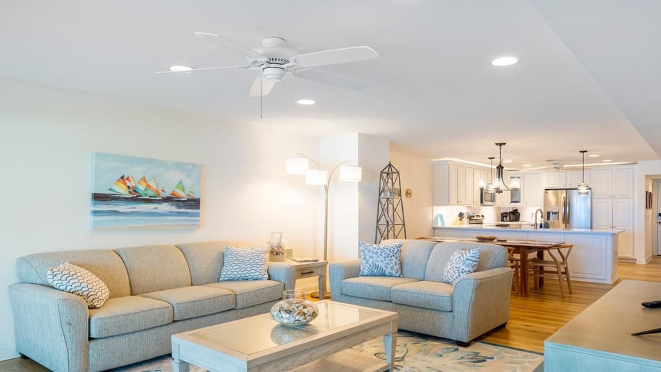 Open-concept living room with beige sofas, patterned throw pillows, coastal artwork, and hardwood floors.
