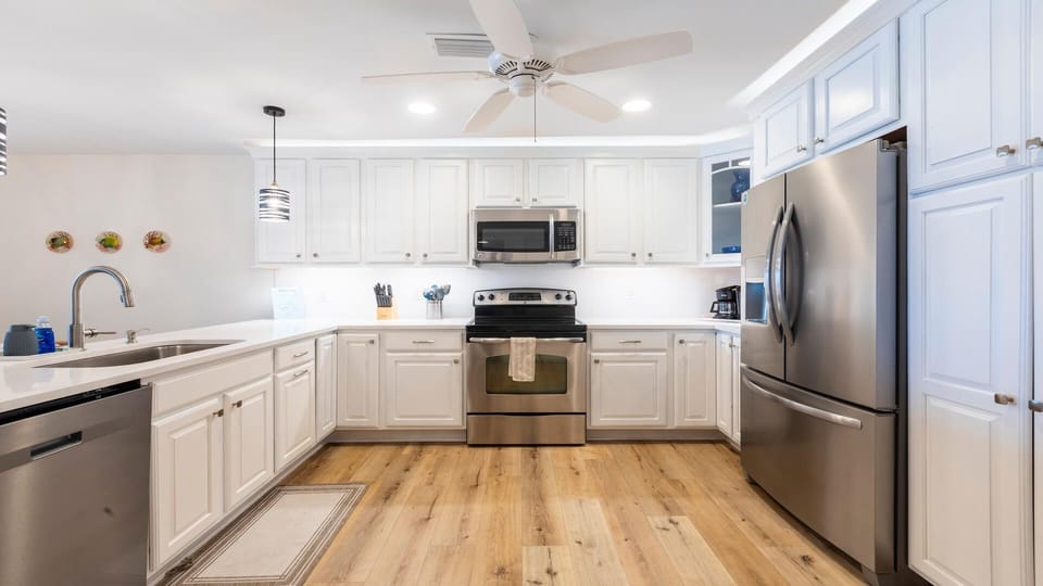 White cabinetry surrounds stainless steel appliances in the kitchen with wood-look flooring and overhead ceiling fan.
