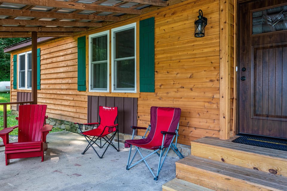 Covered porch with natural wood siding, green shutters, dark wood door, and red outdoor seating on concrete floor.