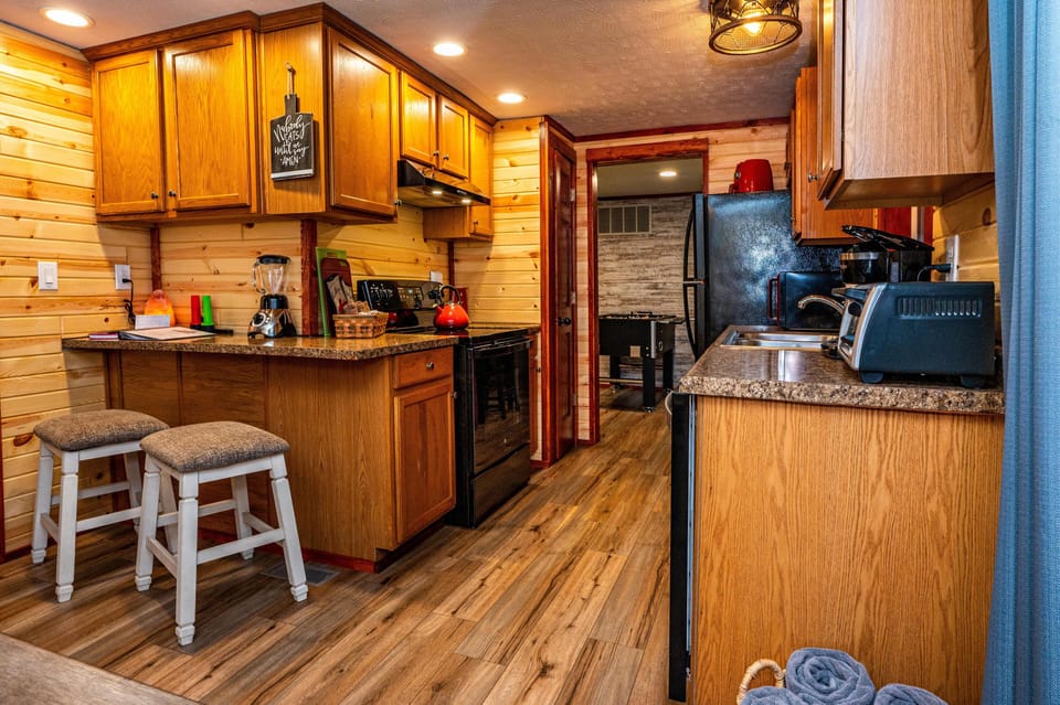 Compact kitchen with oak cabinets, speckled laminate countertops, black appliances, and two cushioned stools.