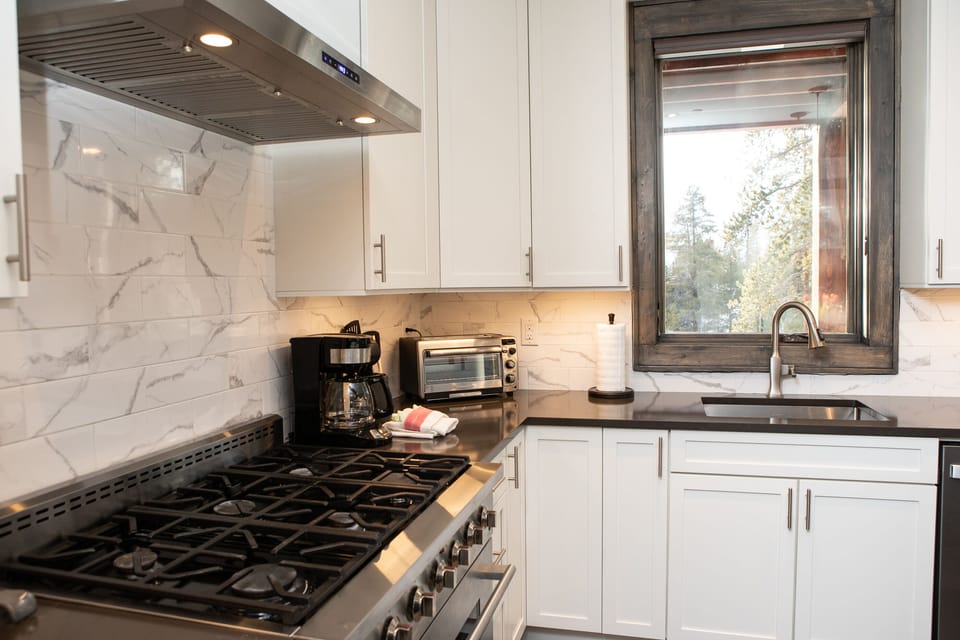 Modern kitchen with stainless steel gas stove, white cabinetry, marble backsplash, coffee maker, toaster oven, and sink beneath a large window overlooking a wooded area.