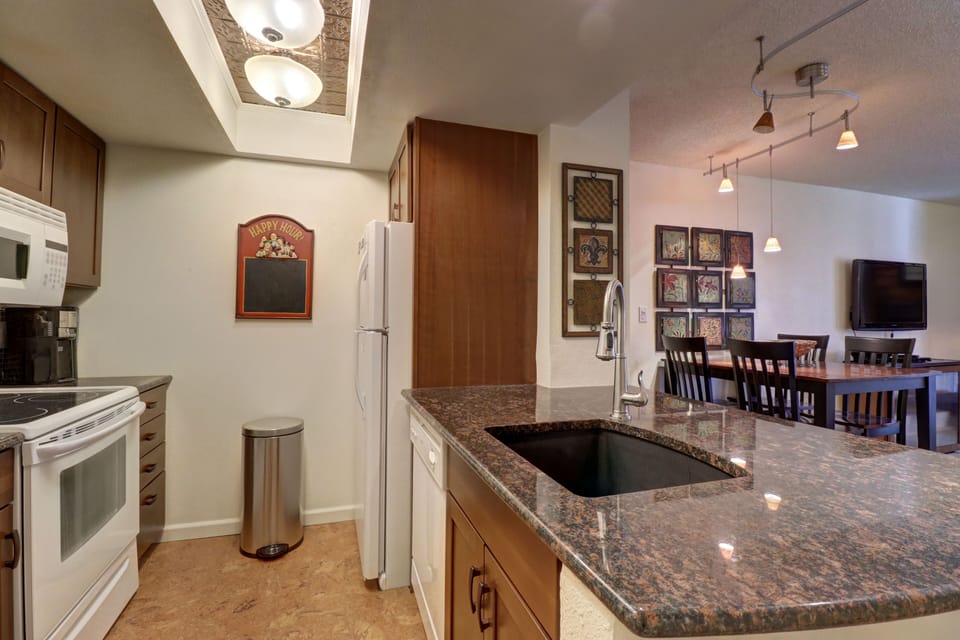 A kitchen and dining area with wooden cabinets, a white refrigerator, stove, microwave, and a granite countertop. There are framed pictures on the wall and a dining table with chairs in the background.