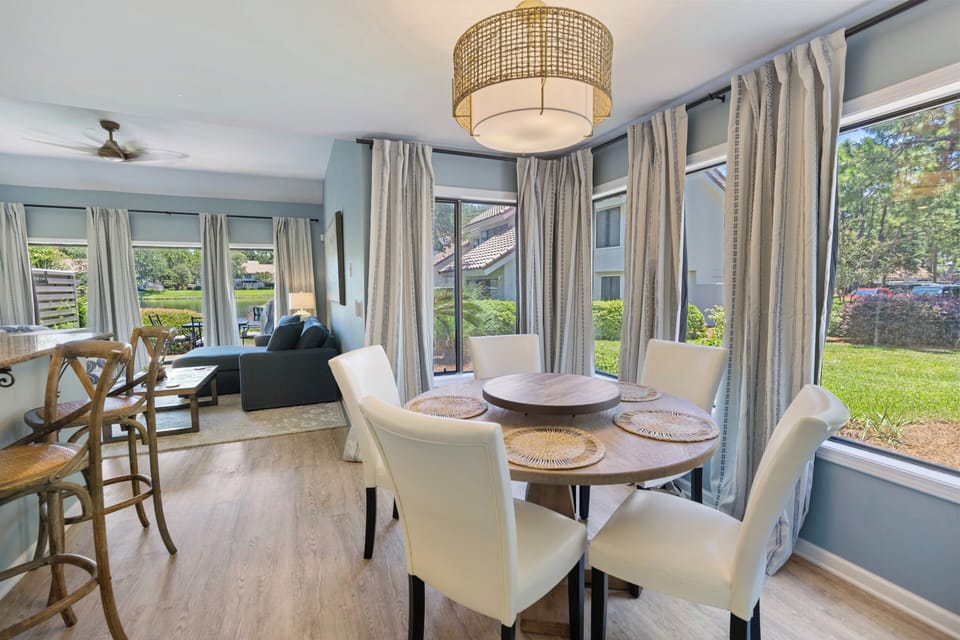 Picture yourself gathering around this inviting dining table, where sunlight streams through expansive windows framing serene garden views. The open layout flows seamlessly to a cozy living space, perfect for relaxed vacation mornings and evenings together