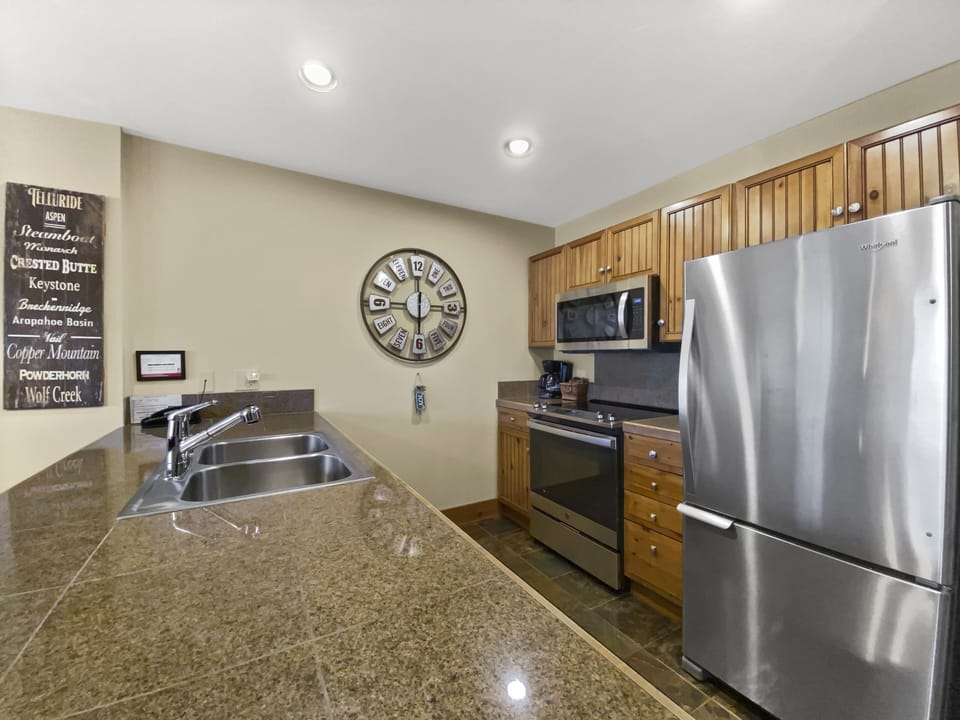 Modern kitchen with granite countertops, stainless steel appliances, wooden cabinets, a wall clock, and a double sink. A framed sign listing names is on the wall.
