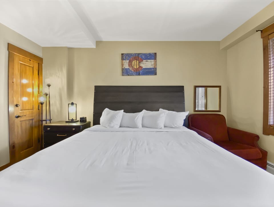 A neatly made bed with white linens in a bedroom. A wooden headboard, nightstand with a lamp, framed mirror, and red armchair are against the walls. A door and wall art are visible.