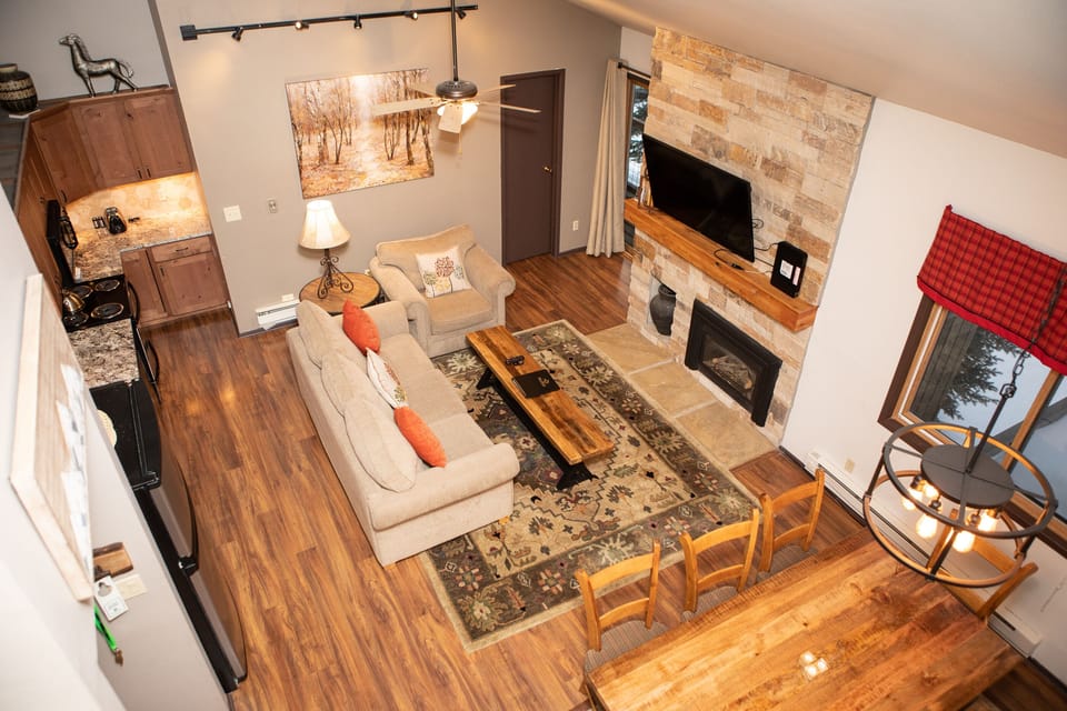 A cozy living area with a sofa, coffee table, stone fireplace, wall-mounted TV, and dining table. The space features hardwood floors, a kitchen, and a large window with a red valance.