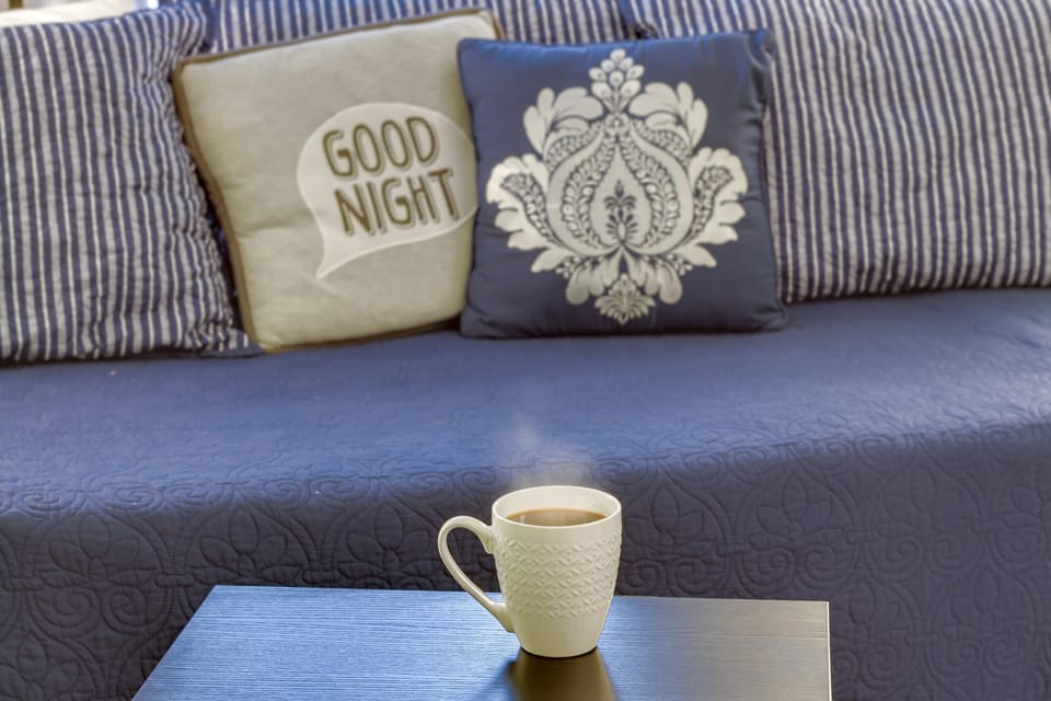 Steaming coffee mug on a dark wood table in front of a navy quilted bedspread and patterned pillows in the bedroom.