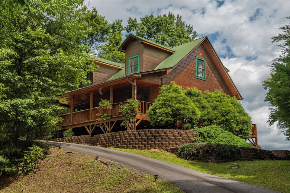 A charming wooden cabin with green-trimmed roof and covered porch nestled in lush mountain greenery, offering a peaceful retreat surrounded by nature.