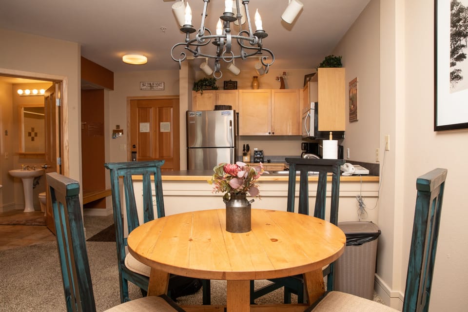A small dining area with a round wooden table and four chairs, a flower vase centerpiece, and a visible kitchen with stainless steel appliances in the background. A bathroom is seen to the left.