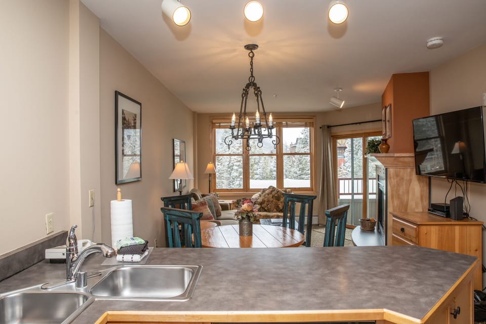 A cozy living and dining area with a round wooden table, chairs, chandelier, TV, and large windows offering a view of snow-covered trees. A kitchen sink and countertop are visible in the foreground.