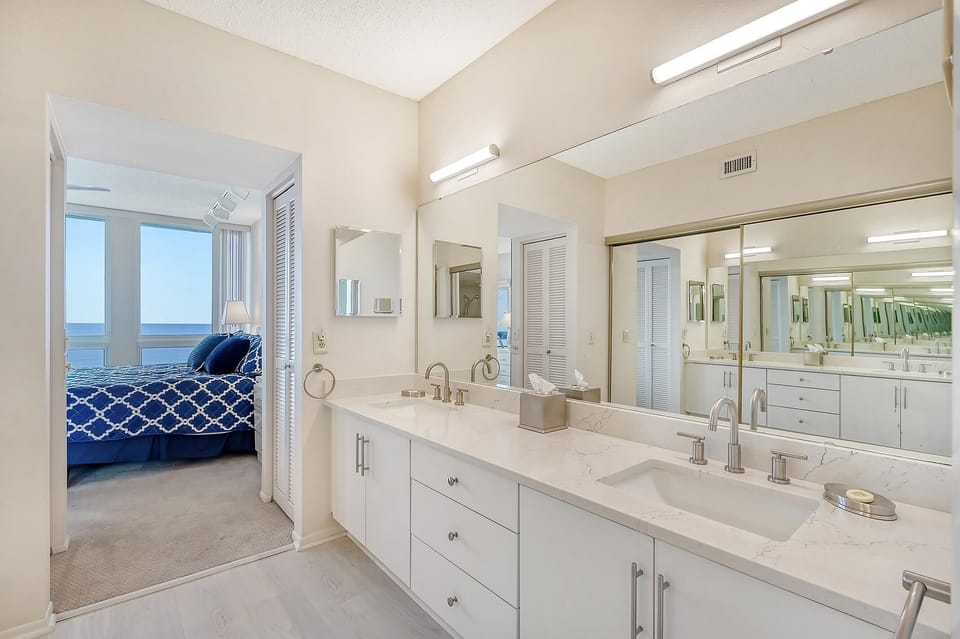 Bathroom vanity with dual undermount sinks, marble-look countertop, and large mirror reflecting the adjoining bedroom.