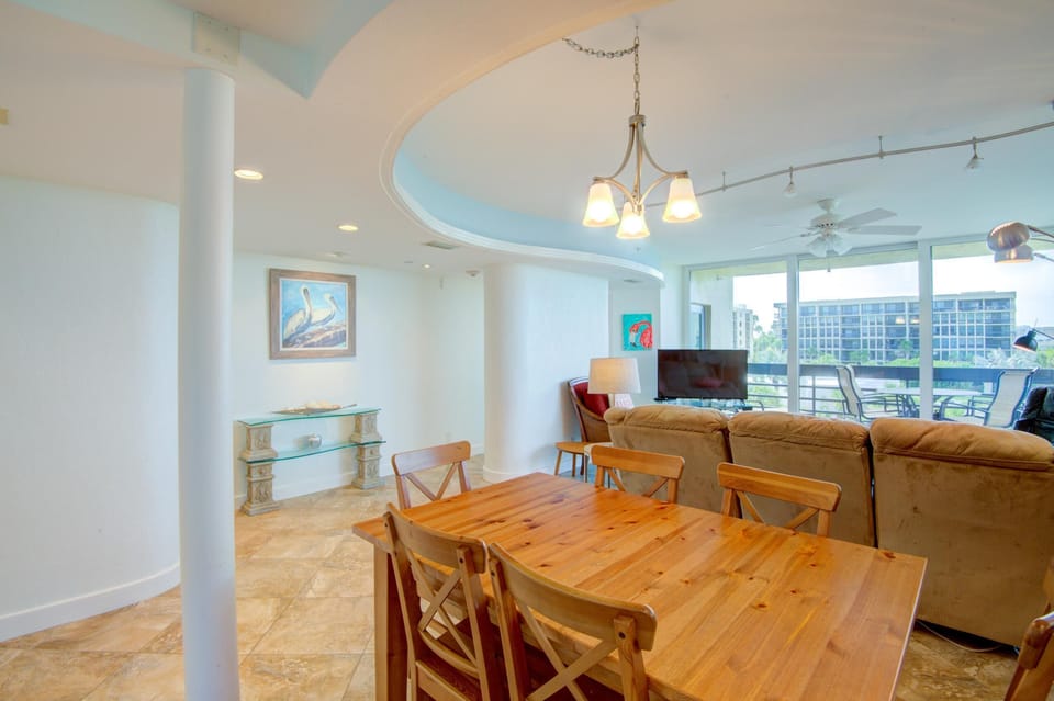 Pine dining table with six chairs beneath a three-light chandelier, adjacent to tiled floors and curved ceiling detail.