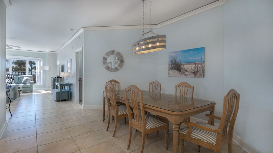 The glass-top table seats six beneath a modern coastal light fixture, with soft neutral tones and crown molding adding a polished finish.