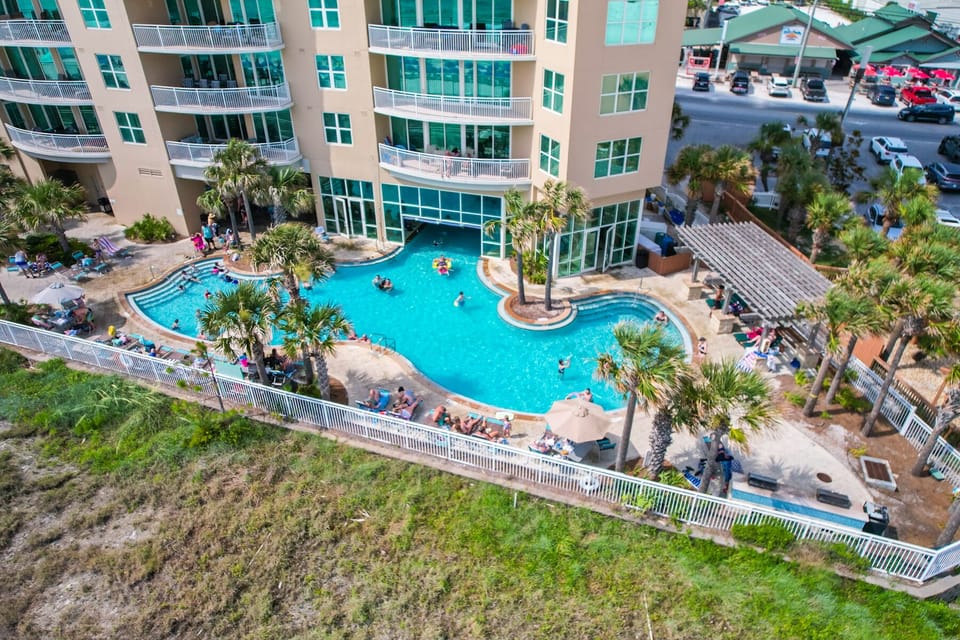 Aerial view of a beachfront resort featuring a curved swimming pool with tropical landscaping and multi-story accommodations.