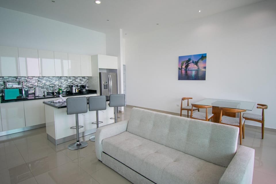 Open-plan living area with light gray sofa, glass dining table, wooden chairs, and white kitchen featuring a metallic backsplash.