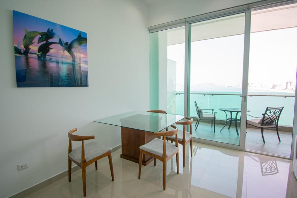 Glass-top dining table surrounded by mid-century wooden chairs beside sliding doors leading to an oceanfront balcony.