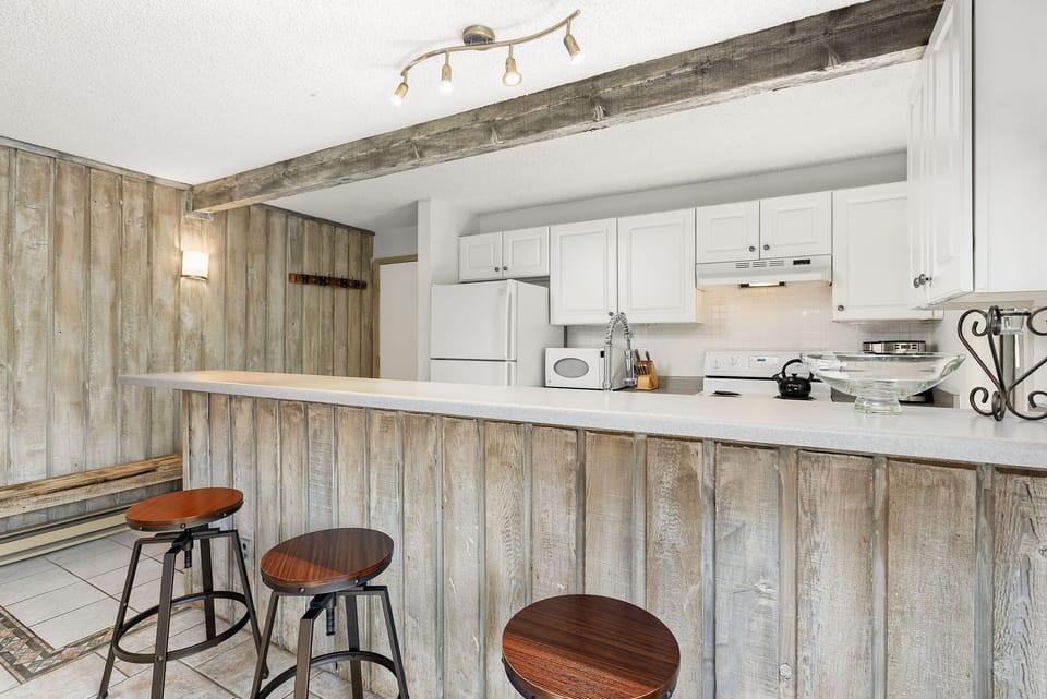 A rustic kitchen features white cabinetry, a white refrigerator, and a breakfast bar with three stools. Exposed wood beams and wood-paneled walls add to the rustic charm.