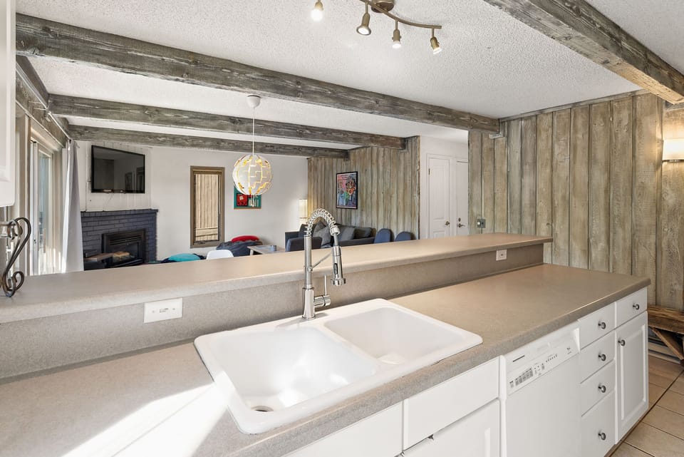 A kitchen with a white double sink and light gray countertops opens to a living area with a fireplace and multiple seating options. The ceiling has exposed wooden beams and a modern light fixture.