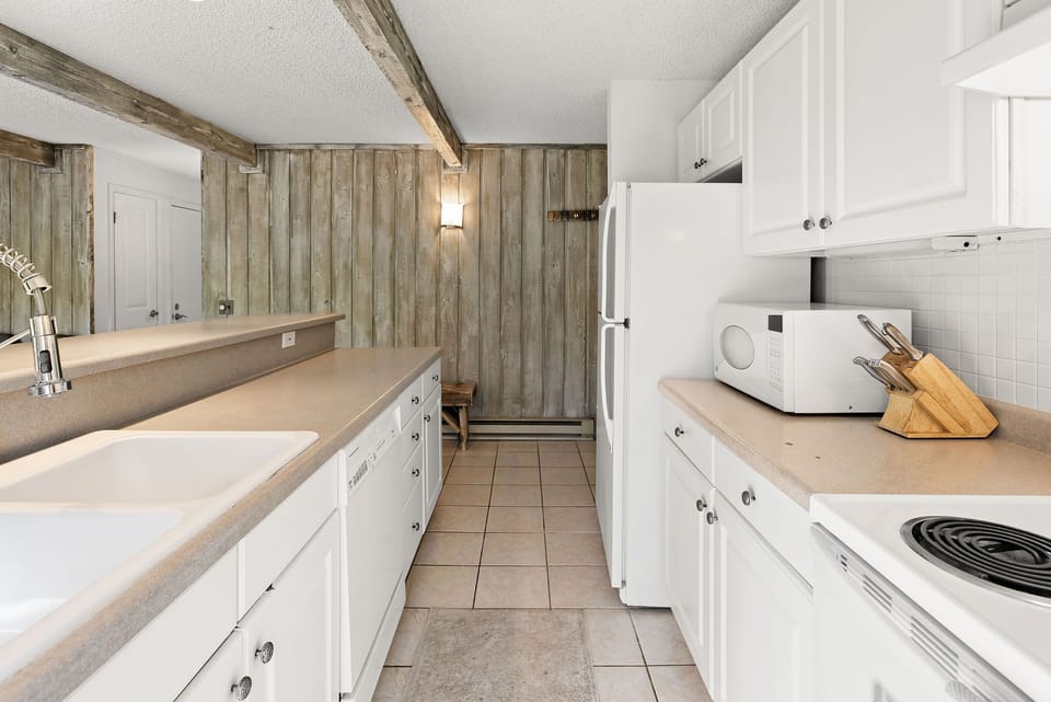 A white modern kitchen features wooden beams, white cabinetry, a refrigerator, a microwave, a sink, a dishwasher, and a countertop with a knife block. Walls are lined with vertical wooden slats.