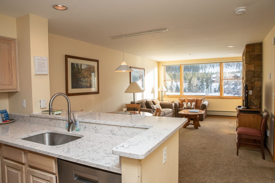 Modern open-plan living space with a kitchen, marble countertops, a living room with sofas, a flat-screen TV, large windows, and a scenic view of a snowy landscape.