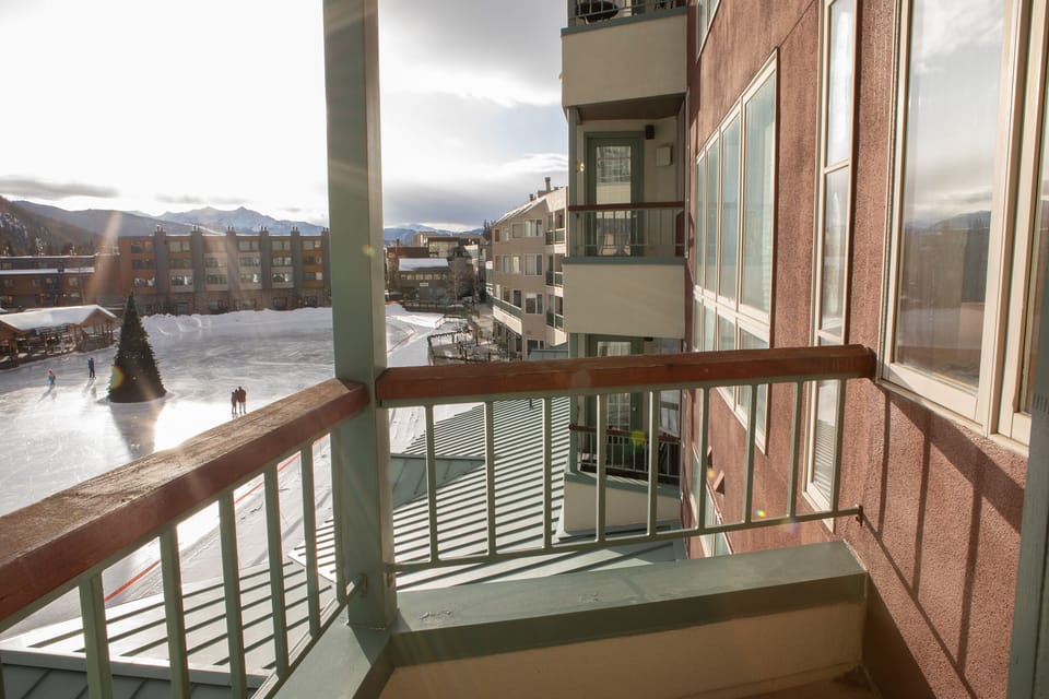 View from a balcony overlooking an ice skating rink and nearby buildings. The sun is shining, casting long shadows, and a few people are visible skating on the rink.