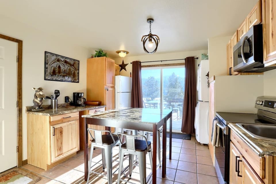 - Sunny eat in kitchen features a granite top island with stools and rustic wood cabinetry
- Full size fridge, coffee maker, and antler style lighting bring cabin warmth
- Sliding glass doors open to views and easy access to the outdoor patio