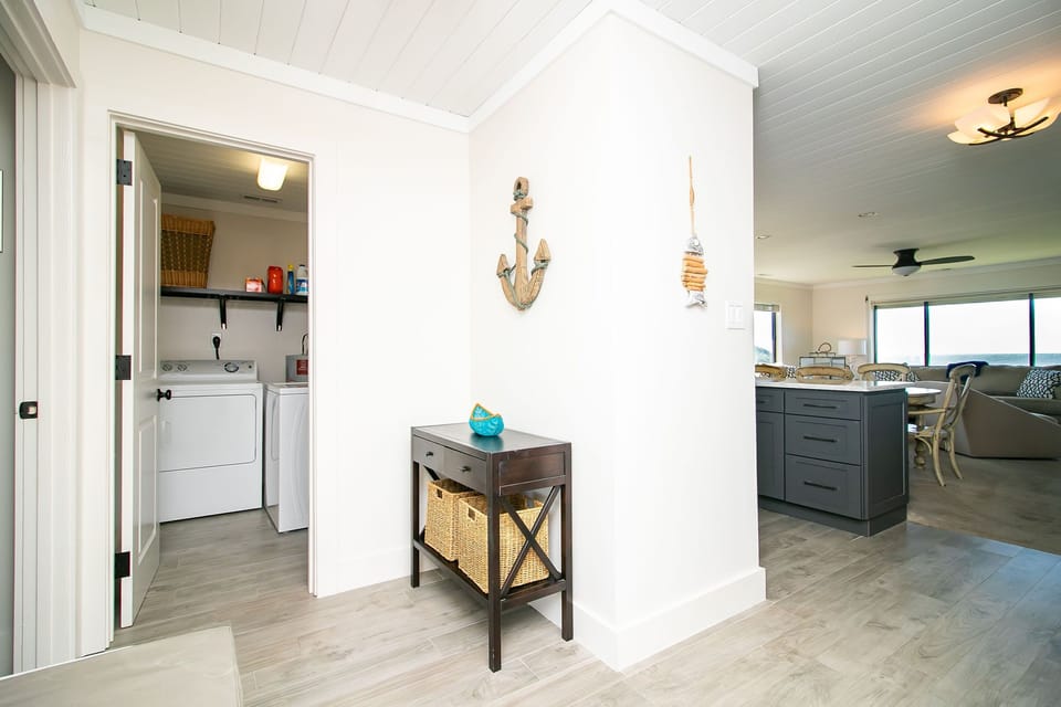 Bright laundry closet with washer, dryer, storage shelf, wicker basket, cleaning supplies, water heater, white door, and wood-look tile floor.