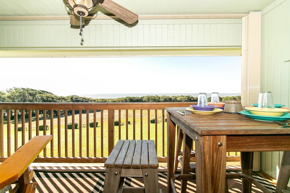 Covered Oak Island deck with wood dining table, bench, ceiling fan, colorful plates, glasses, railing, green lawn, trees, and distant Atlantic Ocean views.