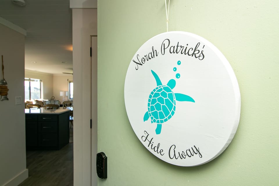 Entry sign with sea turtle logo, pale green wall, door keypad, hallway, and view into a bright Oak Island vacation rental living and kitchen area.
