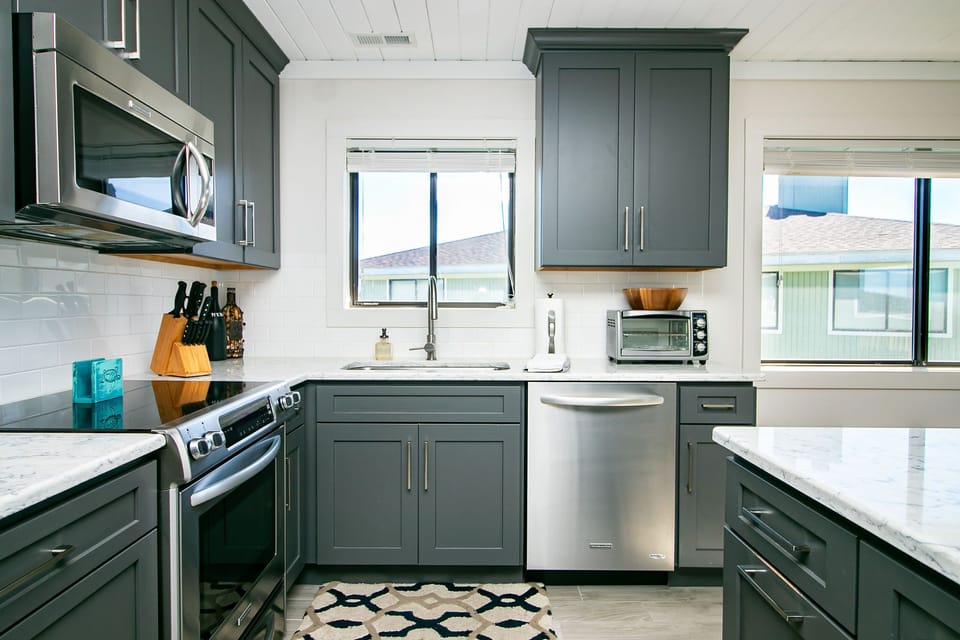 Bright kitchen with gray cabinets, marble-look island, stainless appliances, white backsplash, sink window, toaster oven, and coastal vacation rental style.