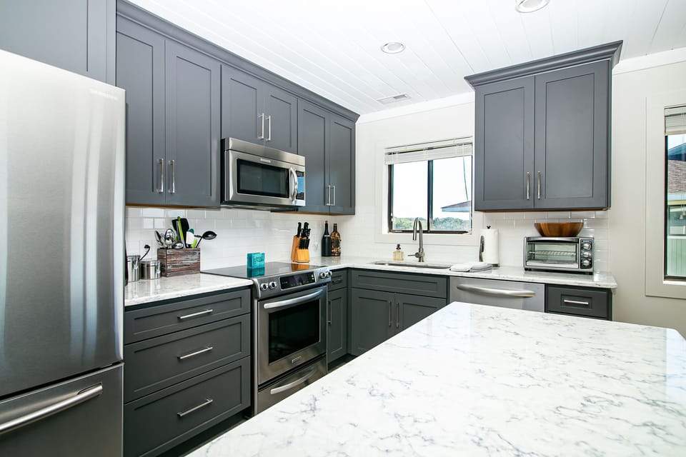 Bright kitchen with gray cabinets, marble-look island, stainless appliances, white backsplash, sink window, toaster oven, and coastal vacation rental style.