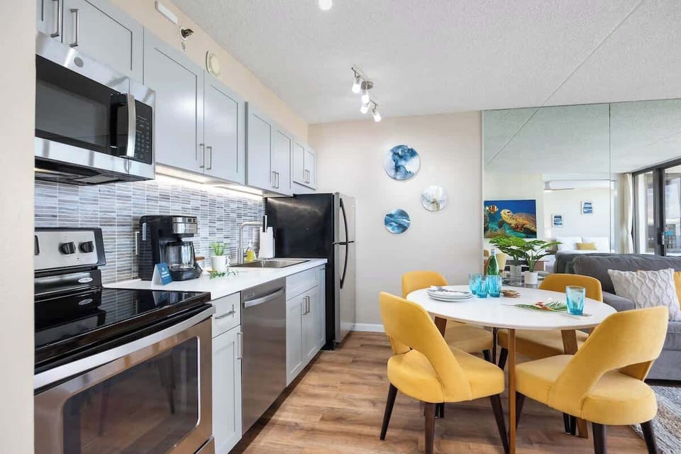 Modern kitchen with stainless steel appliances and a bright dining area featuring yellow chairs.