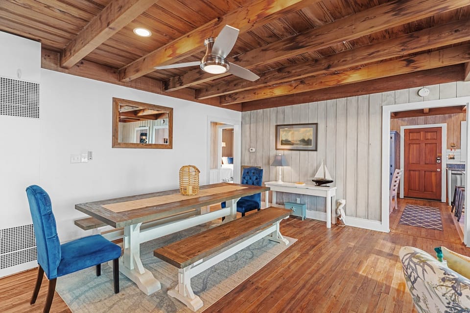 Dining room with a farmhouse table, bench seating, blue chairs, wood floors, cottage wall paneling, and a warm wood beam ceiling.