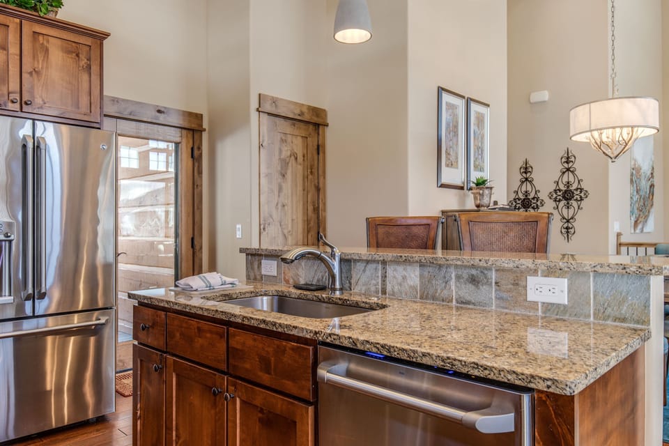 A modern kitchen features granite countertops, a stainless steel refrigerator, a dishwasher, wooden cabinets, and pendant lighting, with a small plant and framed art on the backsplash.