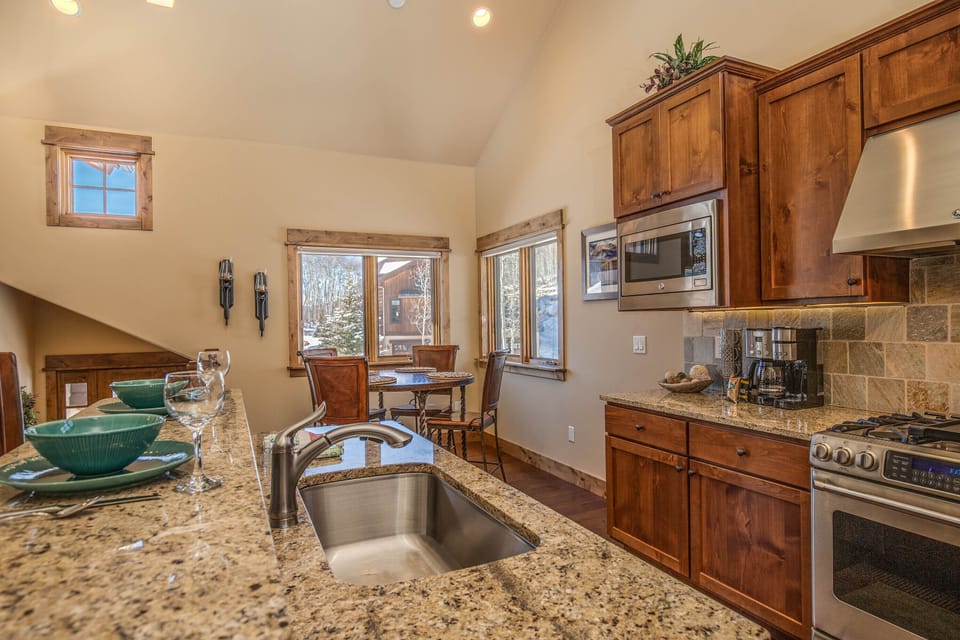 A kitchen with granite countertops, a stainless steel sink, and wooden cabinets. A dining area is visible in the background with a table and chairs near windows. An outdoor view can be seen through the windows.