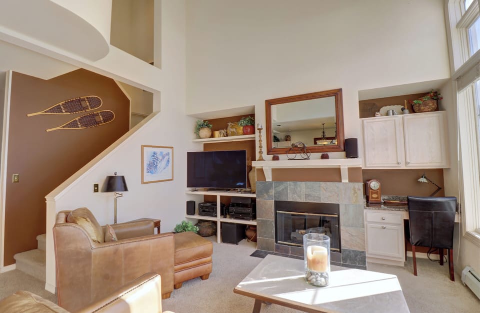 Living room with a cozy armchair, fireplace, TV, and built-in shelves. A small desk with a chair is on the right. Large windows provide ample light.