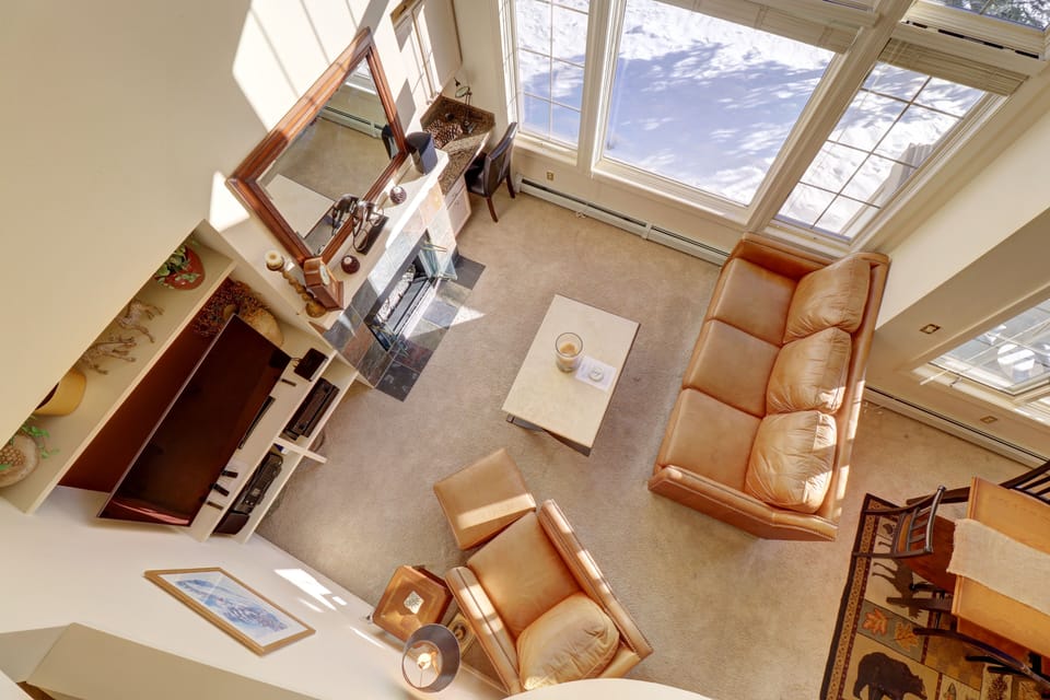 A bright, high ceiling living room with a cozy sofa, a oversized armchair, coffee table, fireplace, and large windows overlooking snowy ski slopes. The room features carpet flooring and a TV.