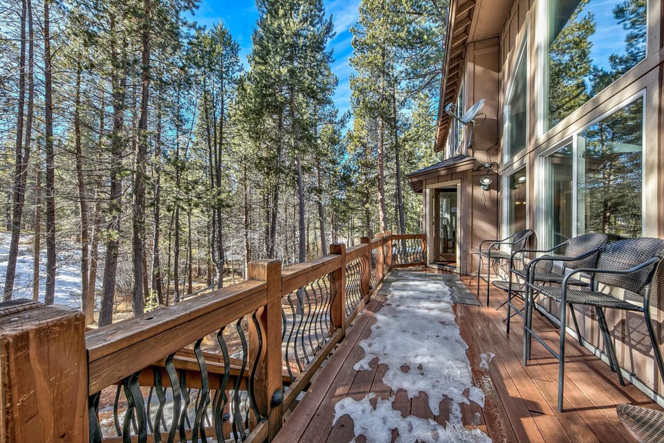 Wood balcony with black metal chairs overlooks a snowy forest through twisted iron railings under a sunny blue sky.