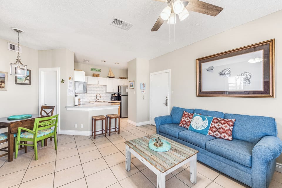 Relax in your bright, coastal-themed living space with comfy blue sofa, open kitchen, and cheerful dining area—perfect for unwinding after beach adventures.