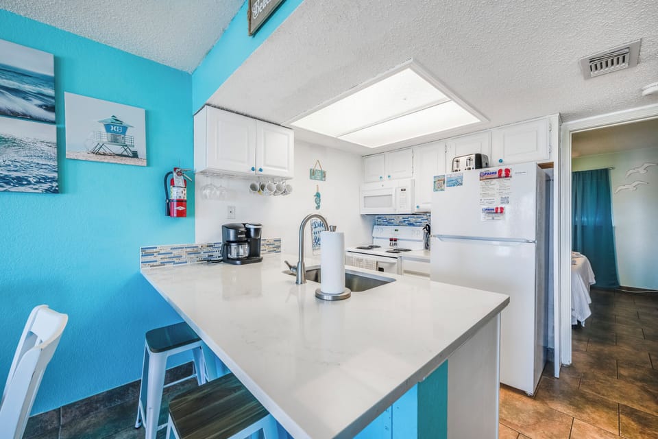 Picture yourself brewing morning coffee in your bright, beach-themed kitchen. White counters, stainless appliances, and coastal blue walls create a cheerful space for preparing meals during your stay.