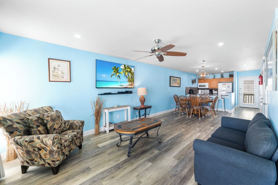 Relax in your spacious open-plan living area with coastal blue walls, comfortable seating, and tropical artwork creating the perfect beach getaway atmosphere.