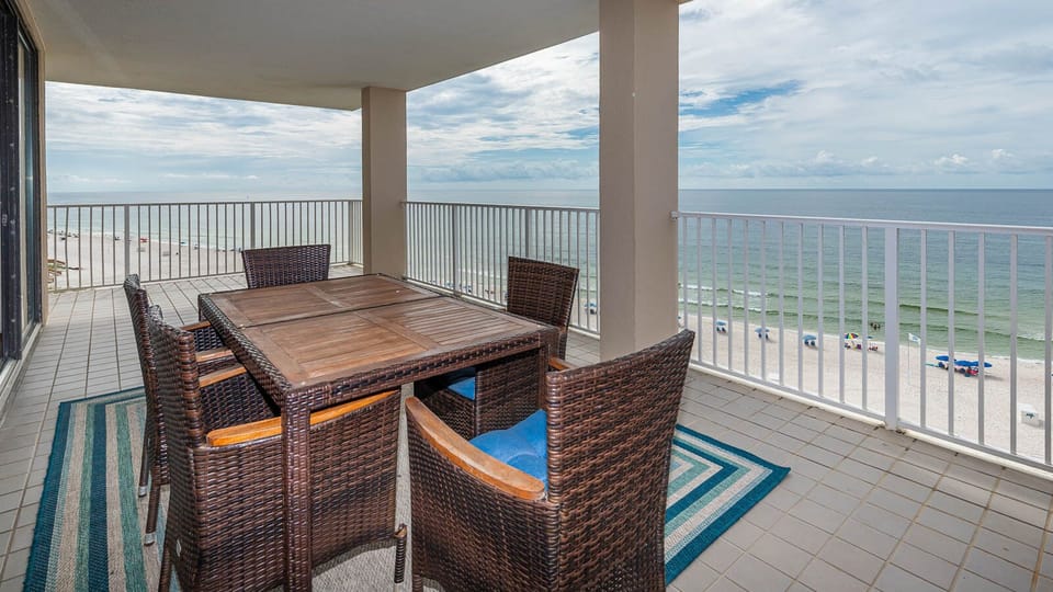 This balcony features a wooden dining table surrounded by wicker chairs, offering a stunning beachfront view