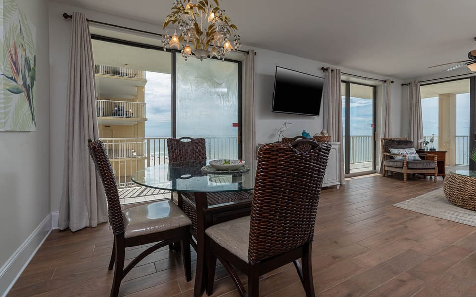 This dining area features a glass table and woven chairs, with a view of the beach and a wall-mounted TV for entertainment