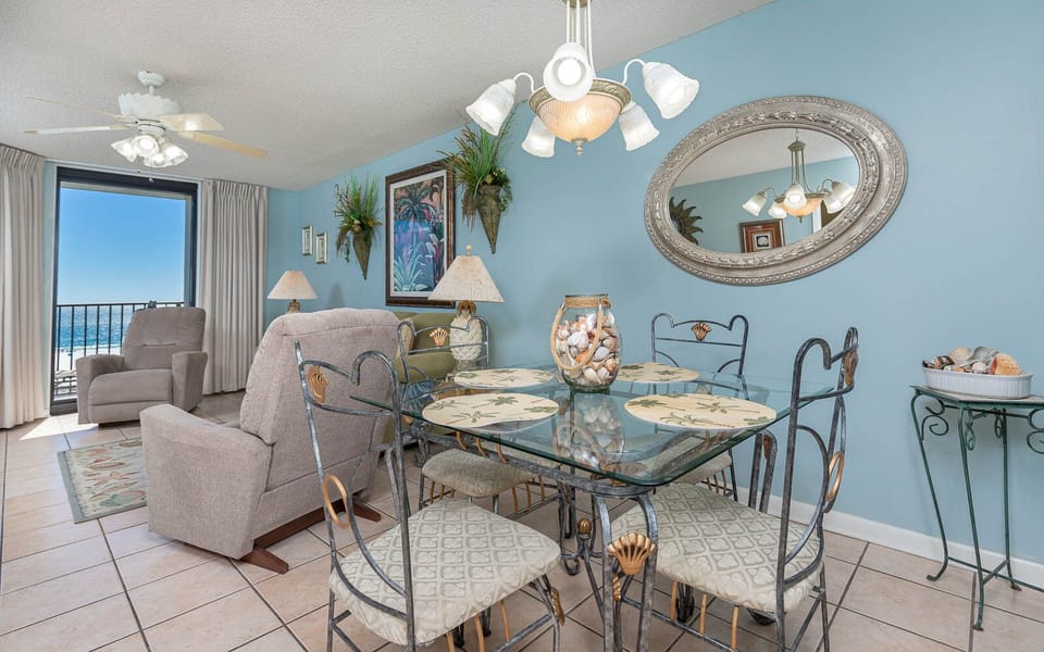 Dining area features a glass table with shell-themed placemats, surrounded by decorative chairs and a view of the gulf