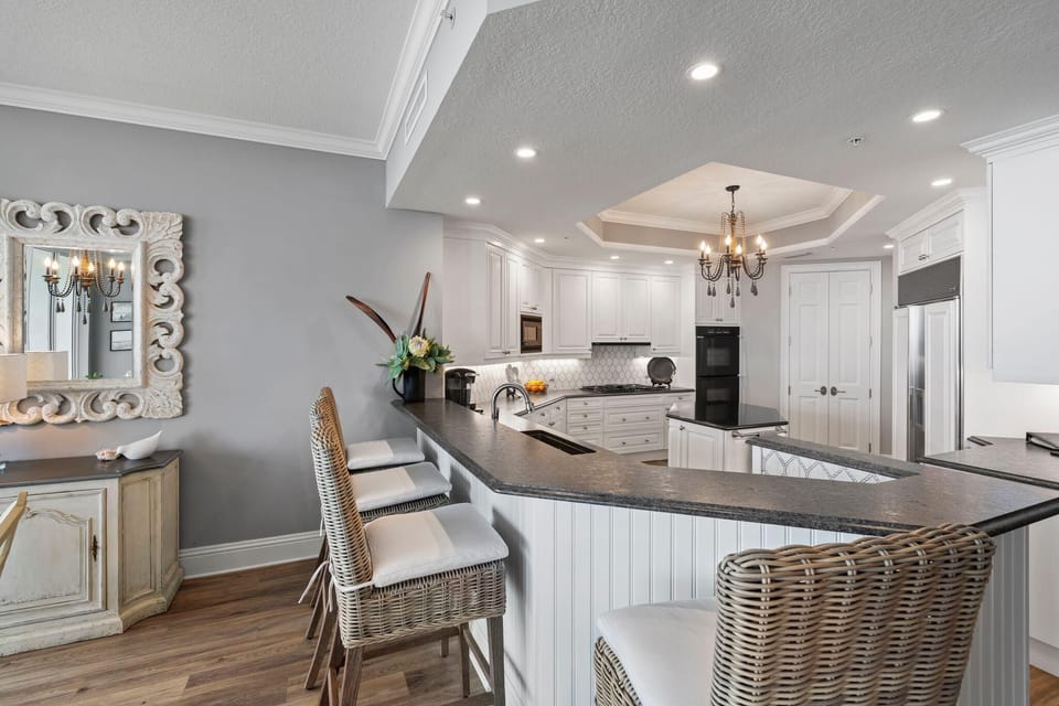 Kitchen with white cabinetry, black granite countertops, wicker bar stools with white cushions, and a wrought-iron chandelier.