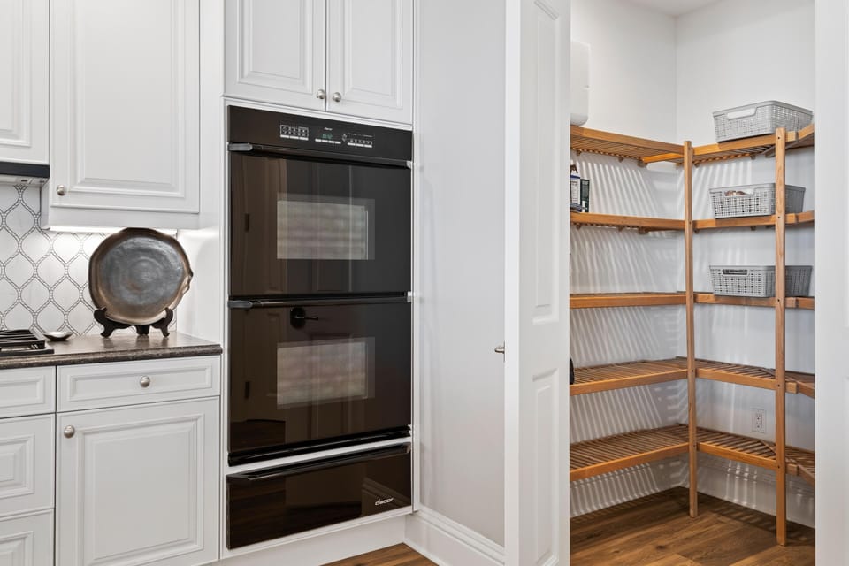 Double black glass ovens are built into white cabinetry beside a pantry with wooden slatted shelves and wicker baskets.